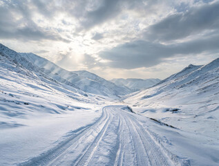 Fototapeta premium A winding snow track cuts through a vast snow-covered mountain valley under a partly cloudy winter sky with sunlight filtering through.