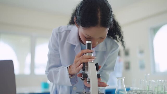 Curly-haired scientist peers through microscope in lab. She wears lab coat, focused on research. Other scientists work nearby in bright lab space. Scientific discovery happens with careful observation