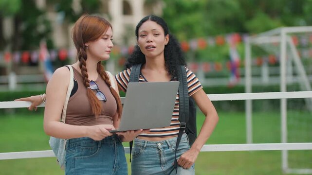 Two students stand outdoors, one holding a laptop. They lean close, eyes focused on the screen. The girl with braids gestures, showing interest in the content. Both smile, suggesting a positive