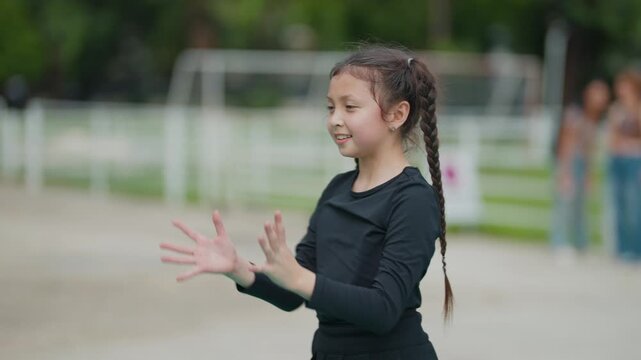 Young girl smiles while playing with a pink ball. Her long braid sways as she runs on a sports field. She wears a black outfit and appears joyful and active