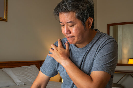 Man holding sore shoulder indoors, bedroom setting with bed and lamp, concept of muscle pain and medical treatment.