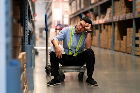 A male warehouse worker wearing a blue shirt is sitting down, appearing distressed or confused amidst the fatigue of warehouse work.