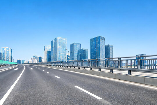 An empty asphalt bridge road leads toward a cluster of modern glass skyscrapers under a clear blue sky.