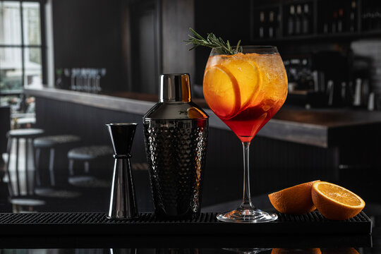 Close-up of an aperol on bar. Next to it is a stylish black shaker with texture and a jigger. In a tall glass there is a bright orange drink with ice and orange slices