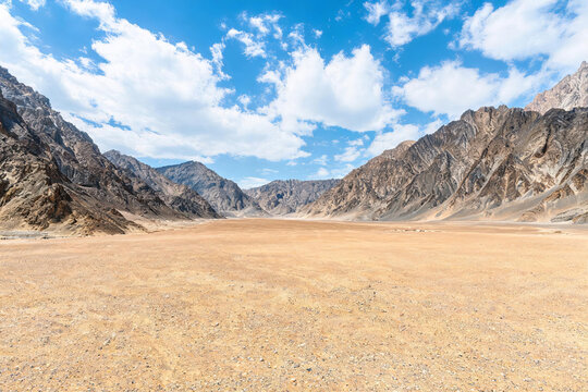 A vast arid valley flanked by rocky mountains under a bright blue sky with scattered white clouds, a barren natural landscape.