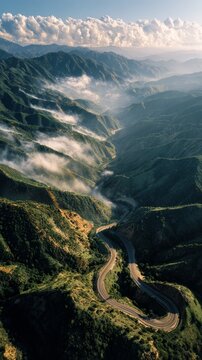 Aerial view of a winding mountain road cutting through lush green valleys with morning mist and clouds