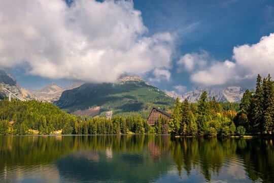 Scenic view of Strbske pleso glacial lake in High Tatras National Park, Slovakia. Vibrant green pine forest and iconic Patria hotel reflecting on water surface under dramatic cloudy sky.