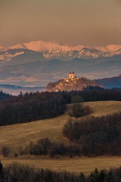 Scenic winter view of Banska Stiavnica unesco calvary church in Slovakia. Red baroque chapels on a hill glowing in warm golden light with snow-capped mountain peaks in distant background.
