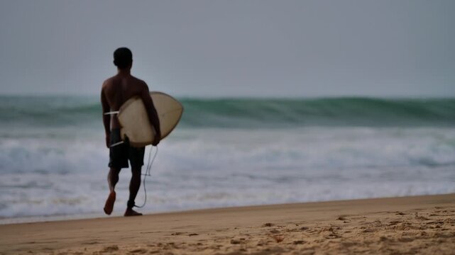Rear view of young male surfer walking along sandy tropical beach holding shortboard surfboard towards ocean waves.