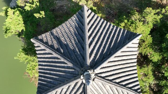 Top down aerial view of Wollyujeong pavilion in Yeongdong South Korea The drone ascends to reveal a traditional hexagonal roof on a rocky cliff by a green river surrounded by lush summer nature