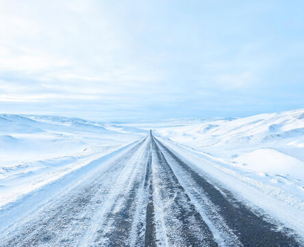 A straight snow-covered road stretches into the horizon, flanked by snow-laden hills under a pale winter sky.