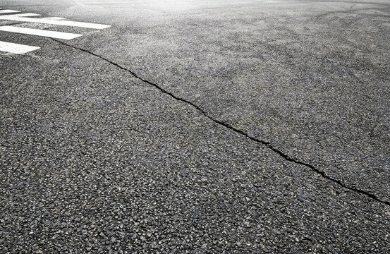 A section of cracked asphalt road with white crosswalk markings, showing pavement wear and infrastructure details.