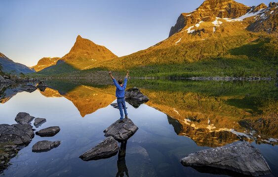 Young man standing on a rock in water, enjoying the landscape, picturesque mountain landscape with Innerdalst&aring;rnet peak in the Innerdalen high valley, mountain peaks reflected in Lake Innerdalsvatna, in the evening light at sunset, Trollheimen Mountain Area, Sunndal, M&oslash;re og Romsdal, Norway
