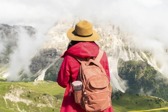 Close up asian female hiker with backpack and straw hat looking at misty mountain peaks deep valley scenic destination Seceda Dolomites South Tyrol Italy. Inspirational ad banner