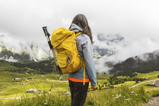 Back view of middle aged gray hair woman hiker yellow backpack overlooking lush green alpine valley misty mountains background Dolomites Alps Italy summer landscape