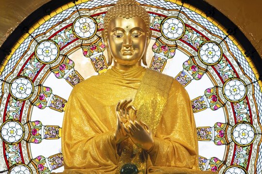 In the main chedi of Wat Phra That Sorn Kaew, the gilded Buddha statue shines out against a luminous glass mandala that fills the space with light and colour. The delicate patterns of the window and the gentle radiance of the robe combine to create an atmosphere of quiet contemplation. This photograph captures the unique harmony of spiritual symbolism, exquisite craftsmanship and meditative tranquillity: the main chedi (Phra That), Wat Phra That Sorn Kaew, Khao Kho Mountains, Phetchabun Province, Isan, Thailand