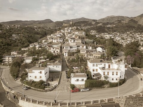 Aerial panoramic view massive stone Gjirokaster Castle on hilltop, surrounded by historic Stone City buildings - Unesco world heritage site. Rugged peaks of Gjere Mountains in Albania. Sepia filter