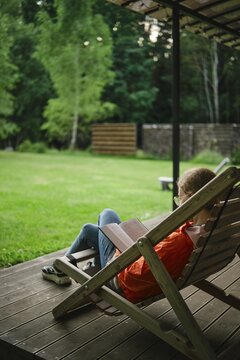 Woman sits on lounge chair in backyard surrounded by grass and trees reading book in afternoon. Calm and quiet atmosphere enhances the enjoyment
