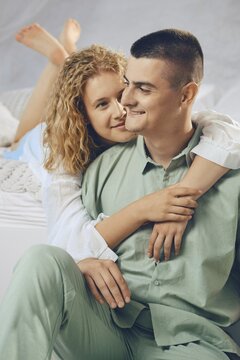 Couple enjoys moment together on bed in bright room. They smile and embrace, showing affection. Natural light comes through window. Relaxed and cozy setting creates warm atmosphere for them