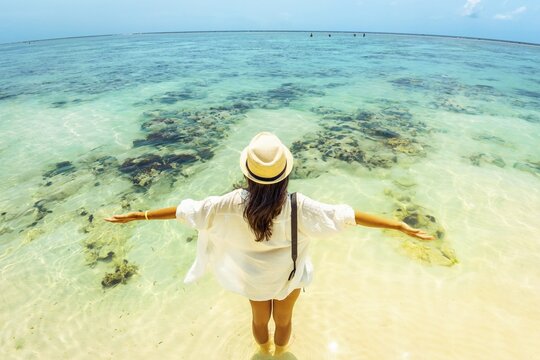 Young carefree woman wear straw hat stands arms wide open hug ocean in shallow, crystal-clear turquoise water on a vast tropical beach. Fish eye lens captures a panoramic view of coral reefs white sand