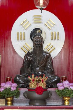 A dark, intricately crafted statue of Laozi (Lao Tzu), the legendary philosopher and founder of Daoism, set against a large Yin-Yang symbol and surrounded by the eight Bagua symbols. Red panels with golden characters and offerings, Chee Chin Khor Temple, Bangkok, Thailand