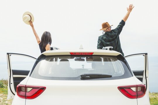 Couple cheers with arms raised while standing by white car overlooking coastal cliff. Bright horizon and open doors evoke sense of freedom and successful road trip journey