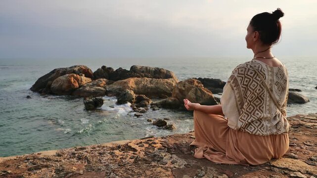 Side view of a serene adult woman sitting in a lotus meditation pose with closed eyes on an ancient stone wall overlooking the ocean bay and rocky shore.