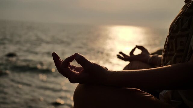 Close-up of a person's hands holding gyana mudra gesture while practicing outdoor yoga meditation by the sea at sunset with warm golden light reflections on the ocean water.