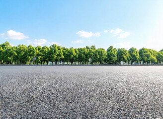 Fototapeta premium A wide smooth asphalt road stretches forward, bordered by a row of lush green trees under a clear blue sky with scattered white clouds, creating a serene outdoor landscape.