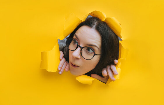 A portrait of a sweet young woman wearing glasses, looking through a hole in yellow paper. A surprised look. Feminine curiosity and gossip. An intellectual.