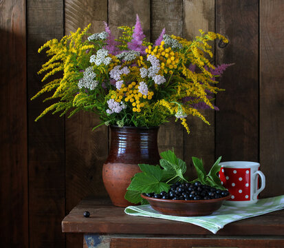 Wildflower bouquet with solidago and yarrow in a ceramic jug sits beside blackcurrants on a wooden table
