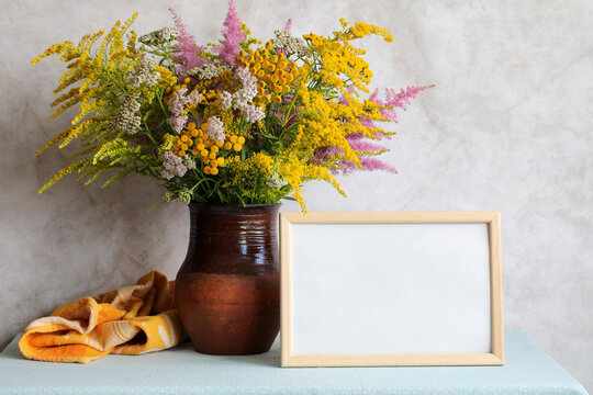 An empty mockup frame and a bouquet of flowers on the table: yarrow and Astilbe, goldenrod and tansy. Summer still life, cottage interior concept.