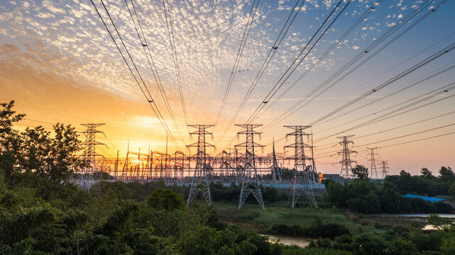 High-voltage power transmission towers and substation facilities stand against a sunset sky, surrounded by natural vegetation and open landscape.