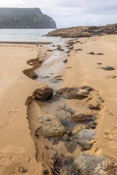 A stream of fresh water from the land flows towards the sea and erodes the sand and exposes rocks on its way across the beach at Steamers Beach in Booderee National Park in New South Wales, Australia.