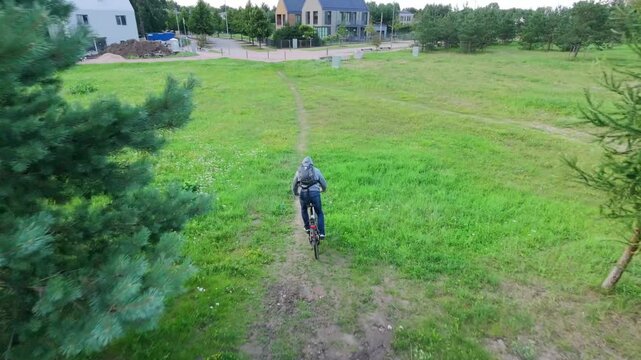 Cyclist moves along a narrow dirt path through a grassy field bordered by young pines. Houses and a paved street appear in the background as the rider follows the track toward the residential area. Th