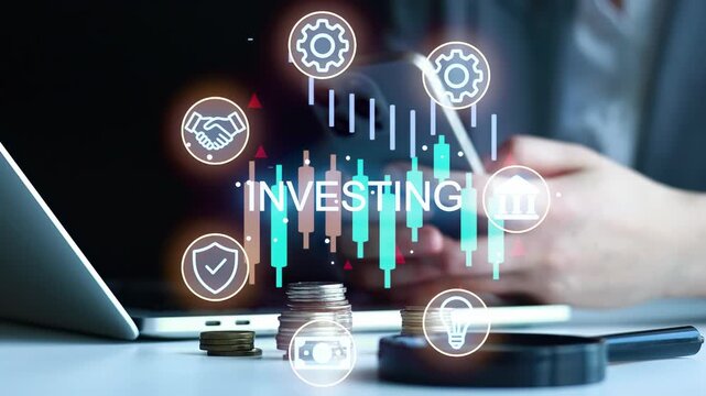 A person is using a laptop and looking at graphs and symbols related to investing. There are stacks of coins on the table. The setting looks like an office.