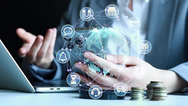 A person is using a smart phone while sitting at a desk with a laptop and piles of coins nearby. Digital icons float above the smart phone. This scene is in an office during the daytime.