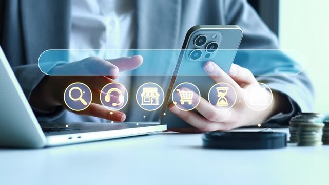 A person is using a smartphone to look for services online. They are seated at a desk with a laptop and some coins around. This scene shows modern technology in use.