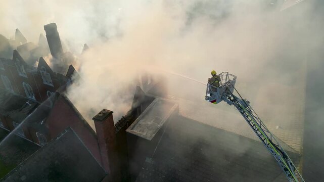Drone shot of a firefighter on an aerial ladder hosing a burning building as thick smoke billows from the collapsed roof