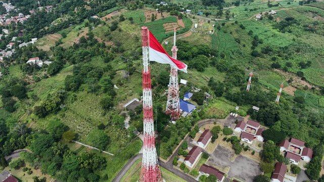 Drone aerial footage of the Indonesian national flag waving atop a telecommunications tower overlooking a lush rural landscape. Symbolizing patriotism, national pride, independence, and modern connect