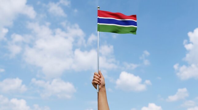Person holding miniature Gambia flag on metal pole against blue sky and clouds in daylight