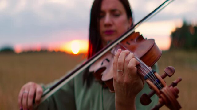 Young woman playing violin outdoors in soft evening light, focusing on delicate fingers and wooden strings against a blurred rural field sunset background