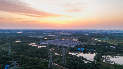 Fototapeta premium An aerial view of an electrical substation and transmission towers set in a rural landscape at sunrise, surrounded by water and greenery.