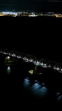 Vertical video. Ufa, Russia. Railway bridge across the Belaya River, steel truss framework illuminated against the dark water and night skyline, Drone footage with city lights on the horizon. Drone f