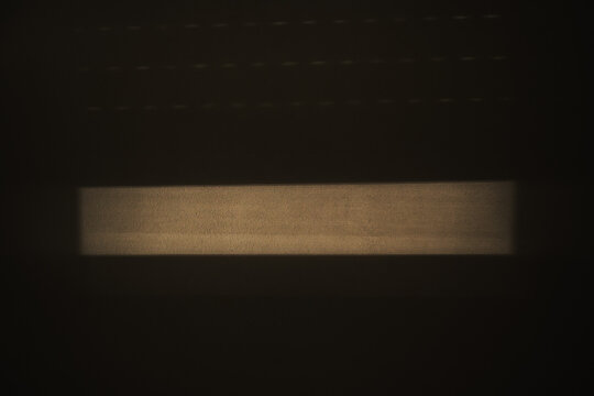 Soft window light casts a beam and shadow across a minimal interior surface, creating an abstract, moody geometric composition with high contrast and subtle texture.