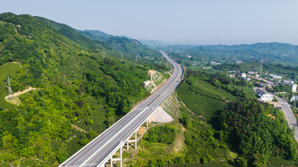 Fototapeta premium A winding highway viaduct cuts through lush green mountainous terrain, with rural settlements and forested hills in the distance.