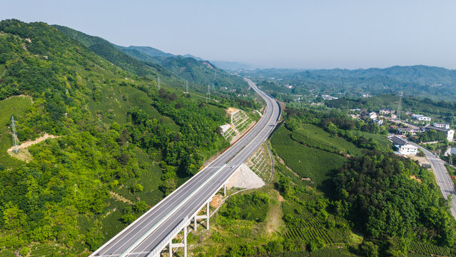 A winding highway viaduct cuts through lush green mountainous terrain, with rural settlements and forested hills in the distance.