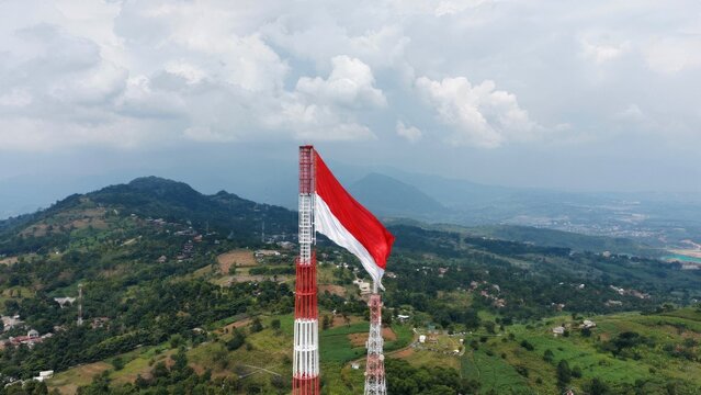 Aerial drone view of the Indonesian national flag waving proudly atop a telecommunications tower overlooking a lush mountain landscape. Symbolizing patriotism, national identity, independence, freedom