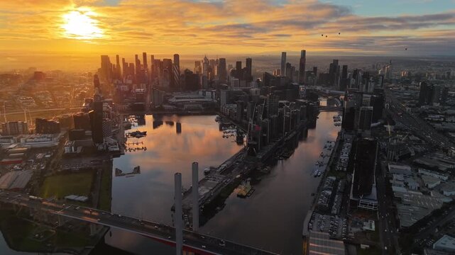 Aerial drone video flying toward Melbourne CBD from above Bolte Bridge at sunrise, showing Docklands harbour, city skyscrapers, golden clouds and reflective water