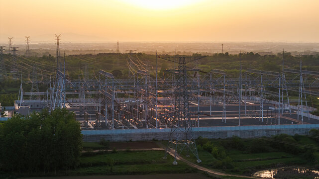 A high-voltage power substation with metal structures situated in a rural landscape during sunset.